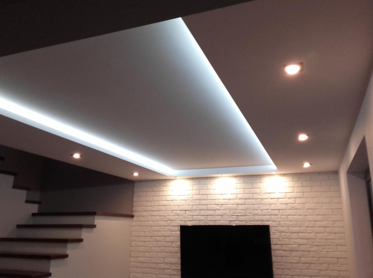 Nowoczesny sufit podwieszany z oświetleniem LED, biała ceglana ściana z czarnym prostokątem, widoczne schody z drewnianymi stopniami.