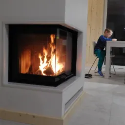 Nowoczesny, narożny kominek z płonącym ogniem, widoczny przez szybę. W tle siedzący przy stole chłopiec.