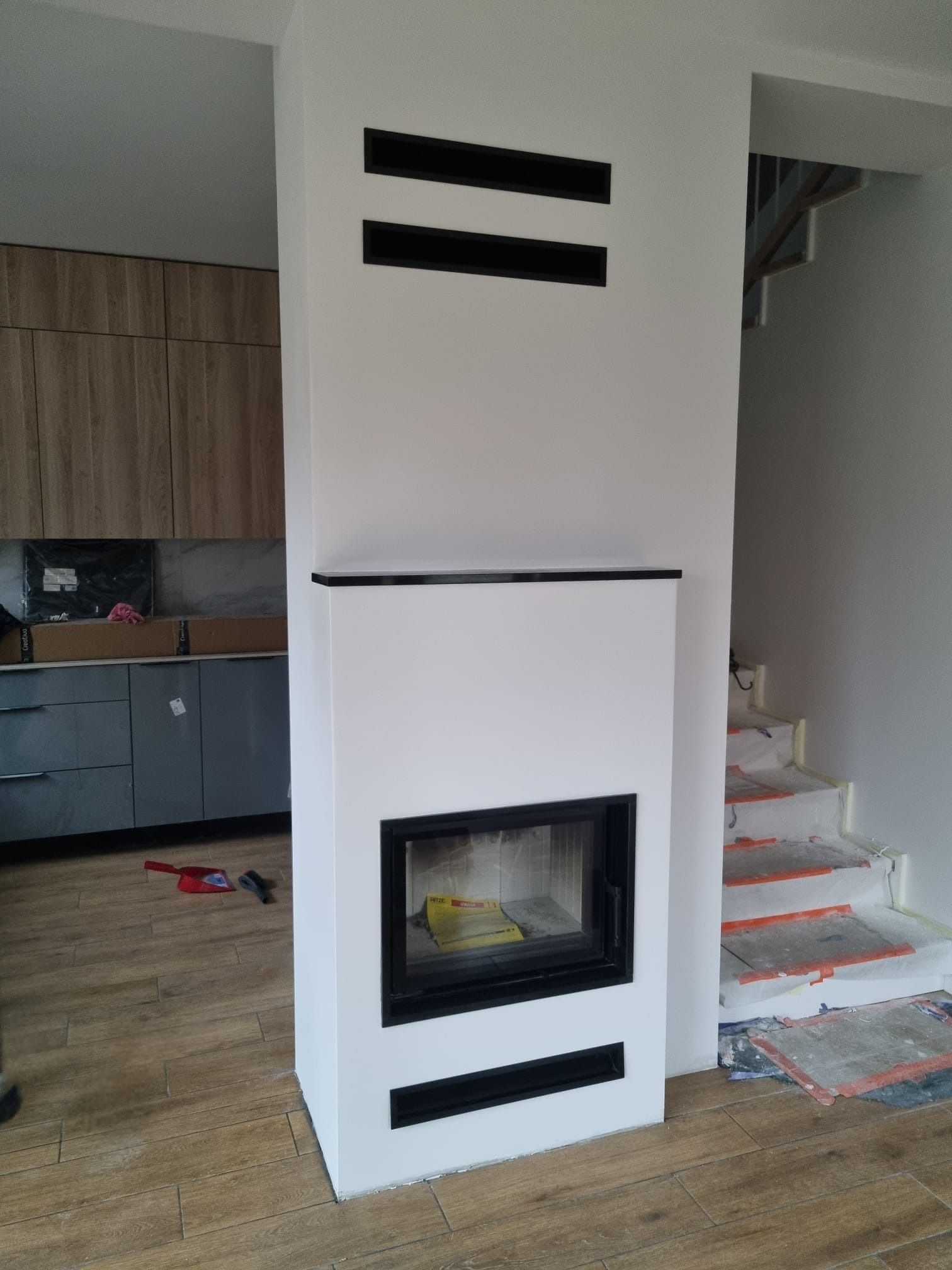 Biały kominek z czarnymi akcentami w nowoczesnym wnętrzu. Widoczne fragmenty kuchni i schodów zabezpieczonych folią malarską. Ujęcie z bliska, detale wykończenia.