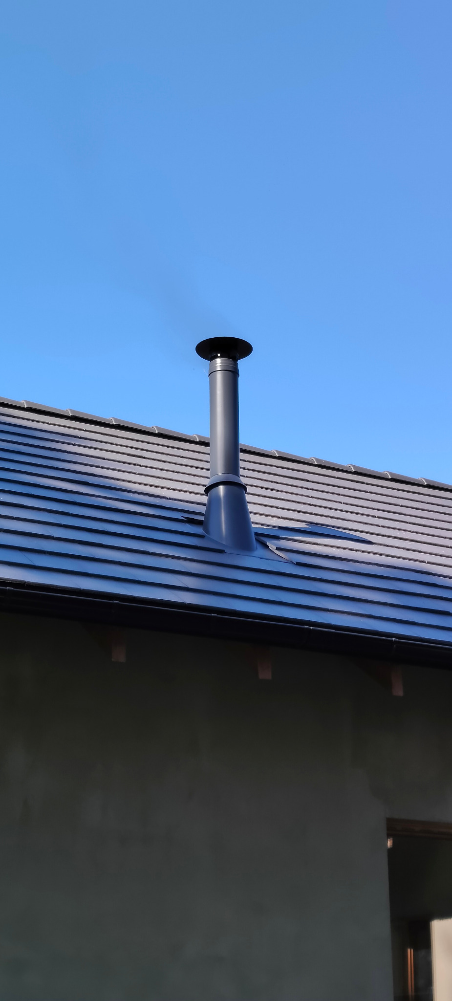 Czarny komin na tle błękitnego nieba i grafitowego dachu z dachówki ceramicznej. Widok z dołu, podkreślający nowoczesny design komina.