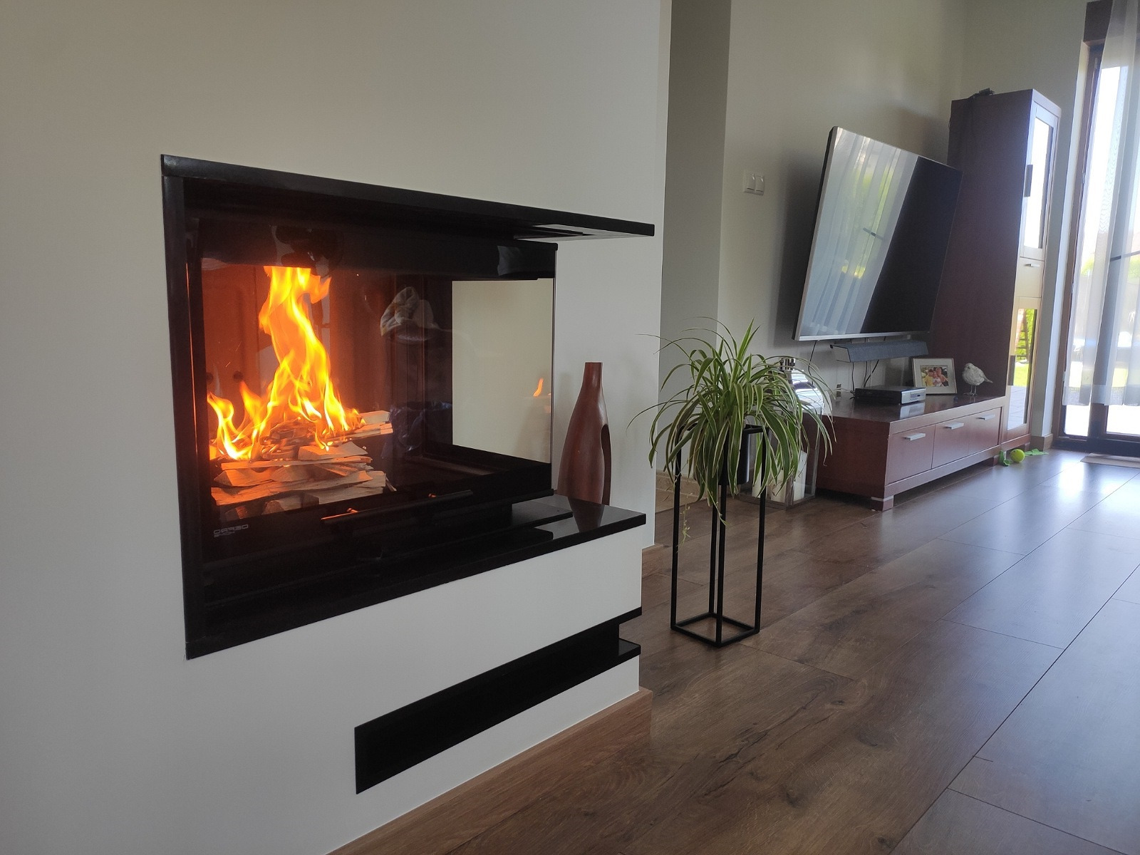 Nowoczesny kominek z czarną obudową i płomieniami, w jasnym salonie z drewnianą podłogą. Obok kominka stoi wysoka, smukła waza i roślina doniczkowa.