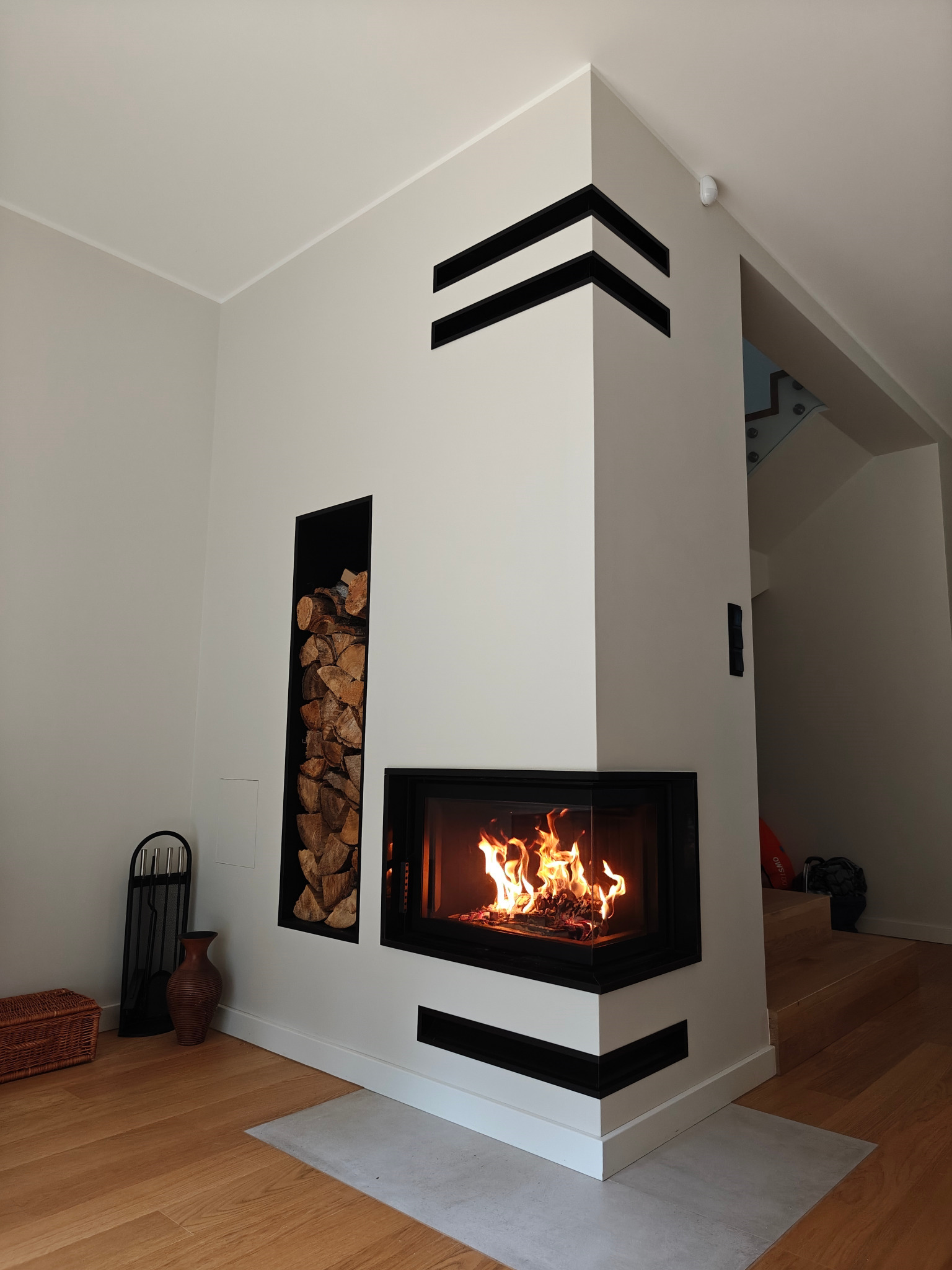 Nowoczesny kominek narożny z czarną obudową i dekoracyjnymi pasami, płomienie w palenisku, obok nisza na drewno. Jasne wnętrze z drewnianą podłogą.