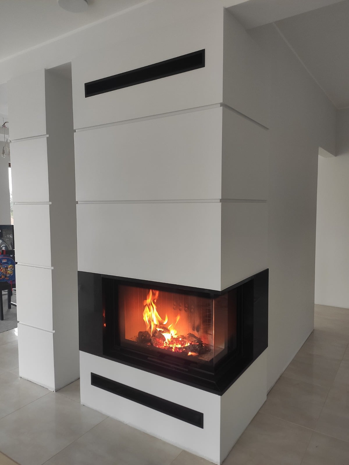 Nowoczesny kominek narożny z czarną obudową i widocznym płomieniem, w jasnym wnętrzu. Minimalistyczny design z poziomymi akcentami. Widok z przodu, lekko z dołu.