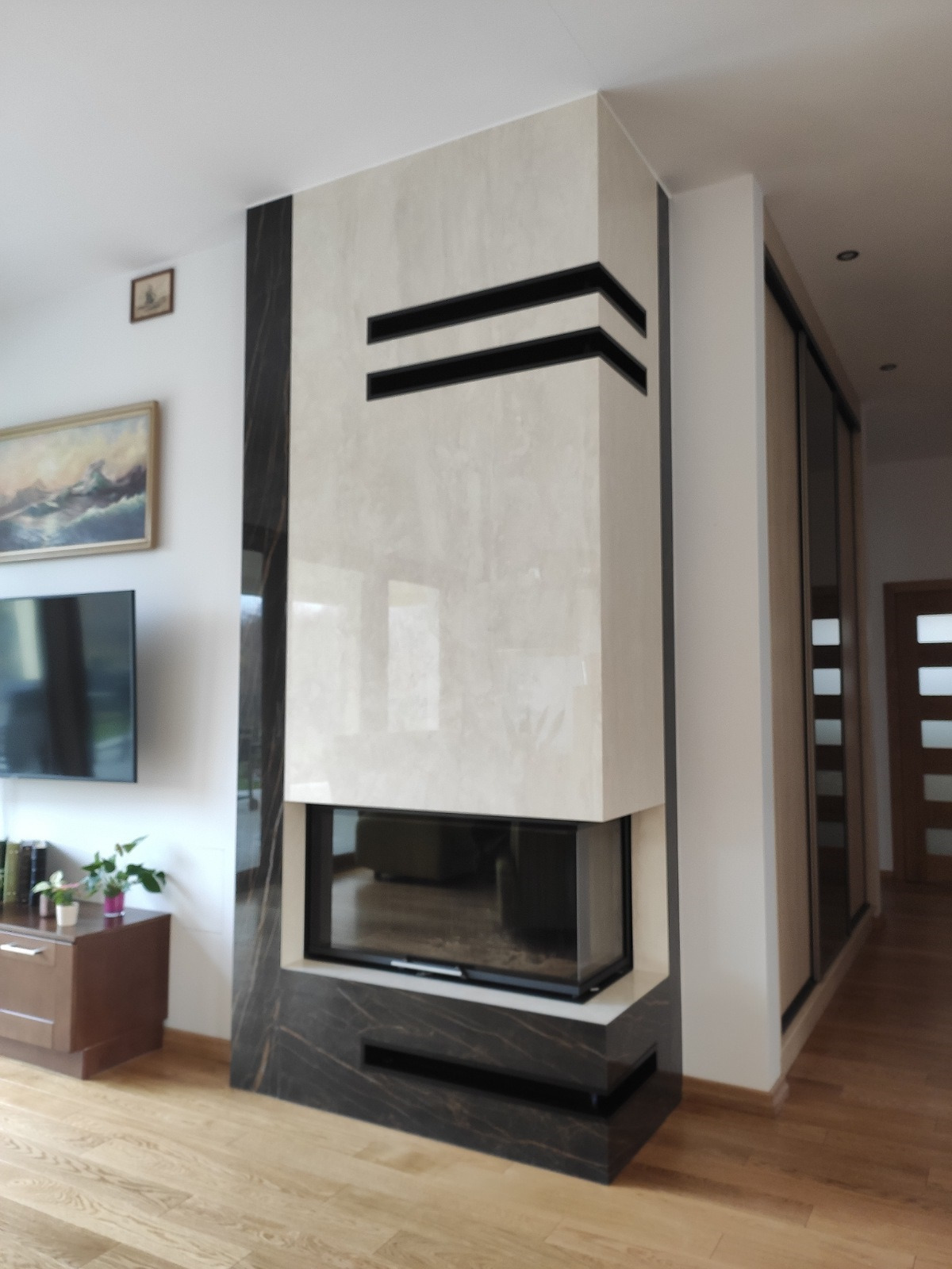 Nowoczesny kominek z obudową z jasnego kamienia i czarnymi akcentami, wkomponowany w jasne wnętrze salonu z drewnianą podłogą i telewizorem na ścianie.