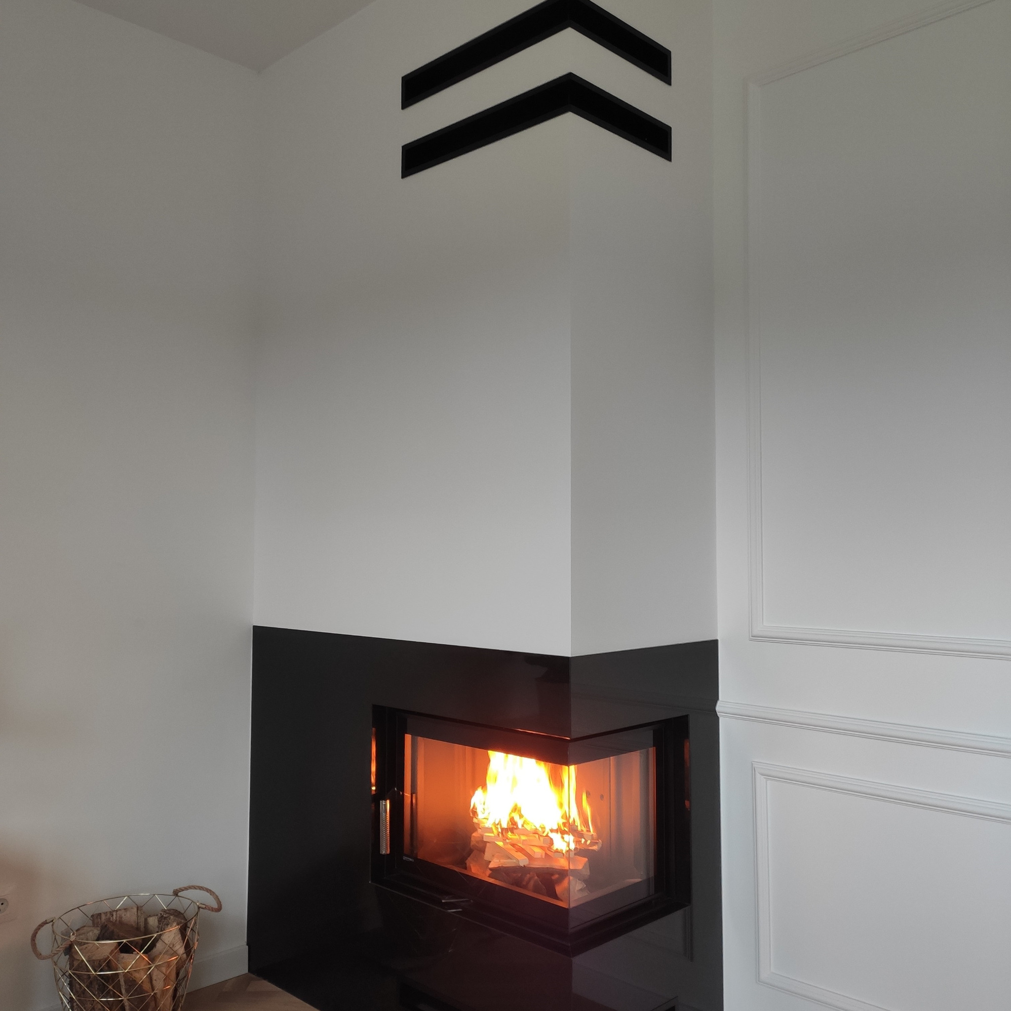 Nowoczesny kominek narożny z czarną obudową i płomieniami, kosz z drewnem obok. Dekoracyjne elementy na ścianie. Minimalistyczny design.