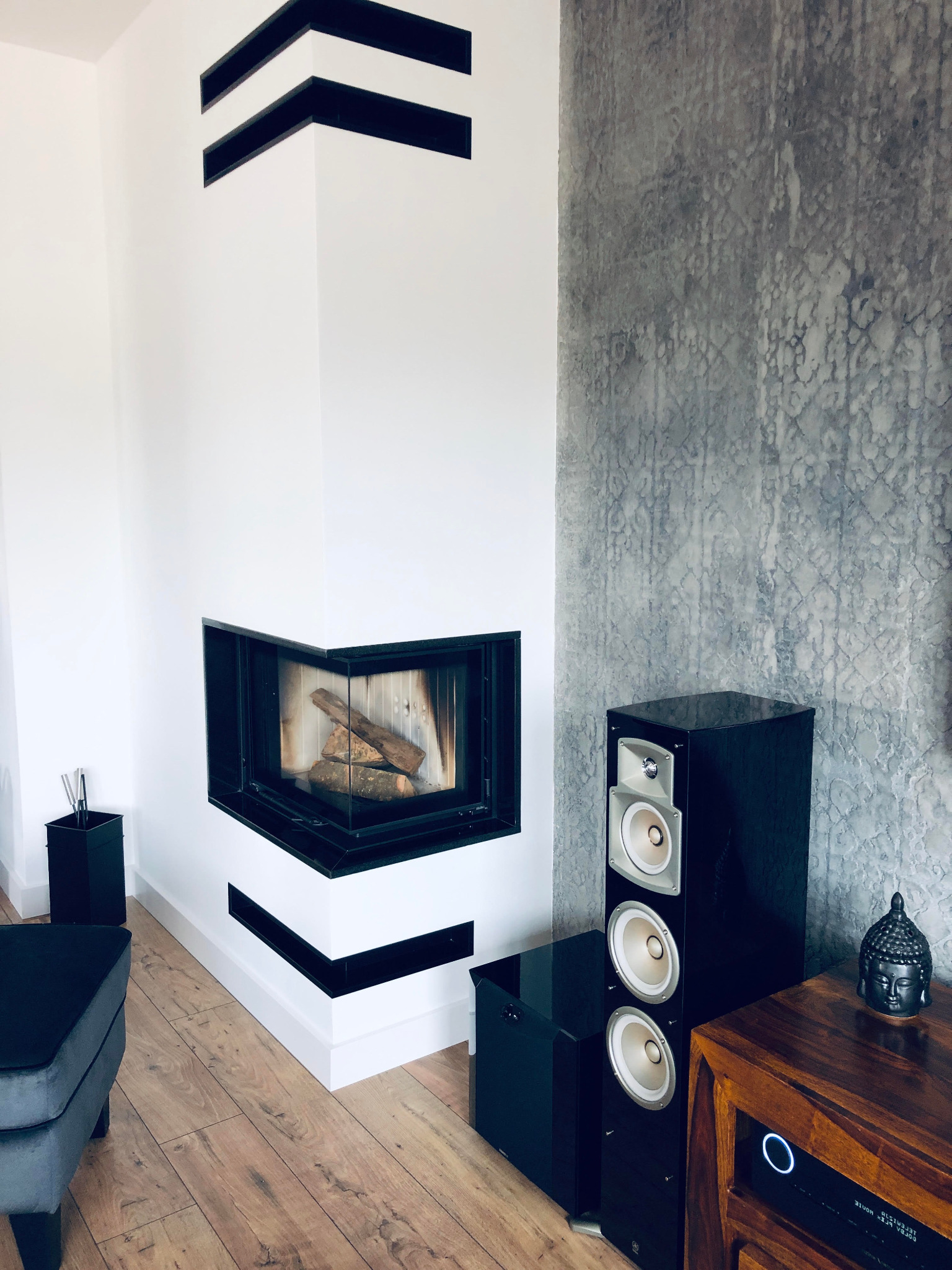 Nowoczesny kominek narożny z czarnymi akcentami, drewnem w palenisku i szarym tłem ściany. Obok głośniki i drewniana komoda z dekoracją.