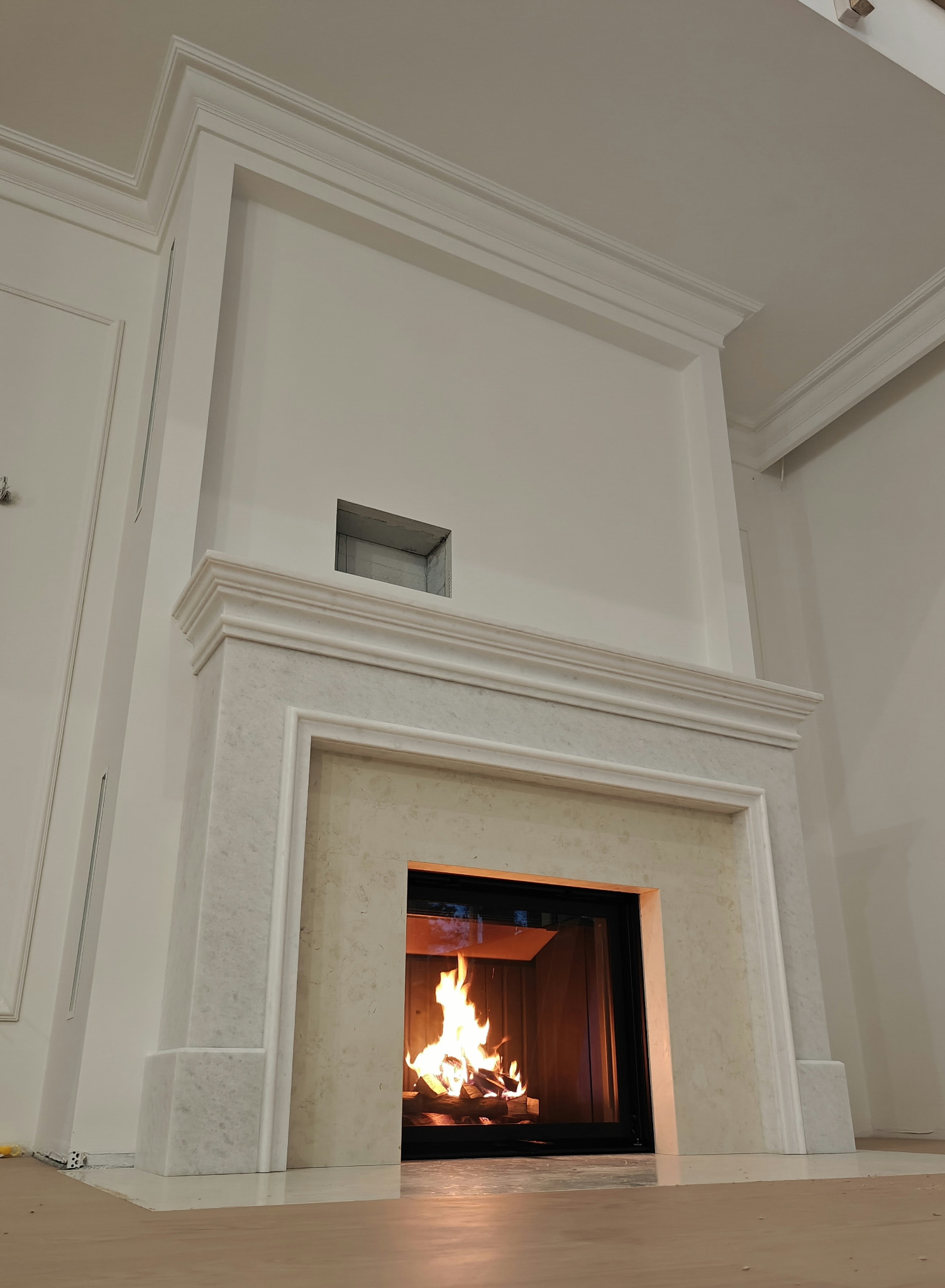 Elegancki kominek z marmurową obudową i paleniskiem, w jasnym wnętrzu. Widoczne detale wykończenia i płomienie. Ujęcie z dołu podkreśla wysokość konstrukcji.