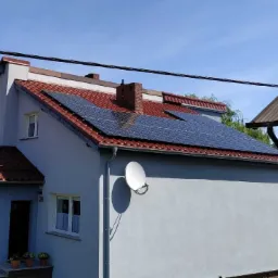 Dom jednorodzinny w Rzepinie z panelami fotowoltaicznymi na czerwonym dachu, antena satelitarna na ścianie bocznej, kwiaty w doniczkach przy wejściu.