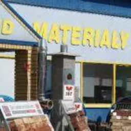 Fasada budynku z żółtym napisem 'EL-BUD MATERIAŁY BUDOWLANE', ekspozycja płytek elewacyjnych i kominów, drabina oparta o ścianę, widoczne okna i drzwi wejściowe.