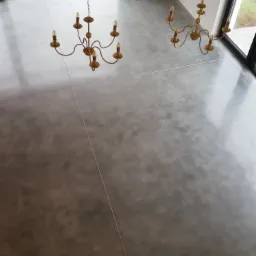 Szara, gładka posadzka przemysłowa z widocznymi śladami polerowania, oświetlona naturalnym światłem z dużych okien i uzupełniona dwoma dekoracyjnymi, złotymi żyrandolami.