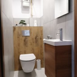 LEVEL - Nowoczesna łazienka z podwieszaną toaletą, umywalką nablatową na drewnianej szafce, szarymi płytkami i drewnianą ścianą za toaletą, na której zamontowano przycisk spłukujący.