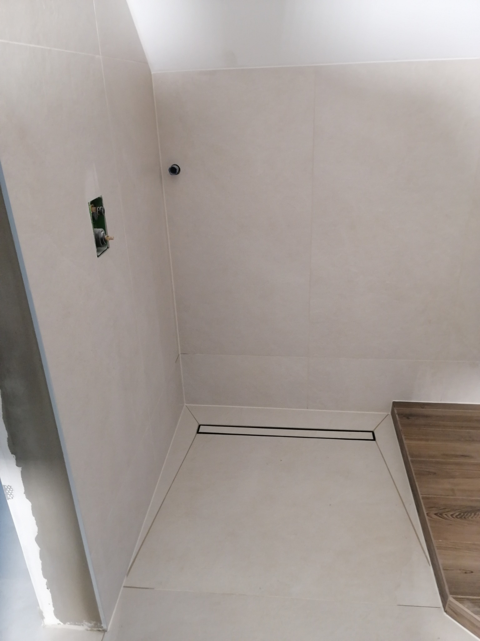 Wykończony brodzik z odpływem liniowym w jasnych płytkach, widoczna instalacja wodna w ścianie i fragment drewnopodobnej obudowy.