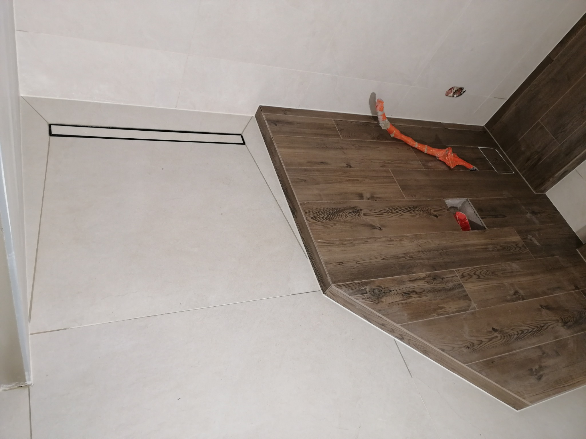 Fragment łazienki z wykończoną strefą prysznicową: białe płytki podłogowe i ścienne sąsiadują z drewnopodobną okładziną podłogową, widoczny liniowy odpływ prysznicowy i wystające rury w folii...