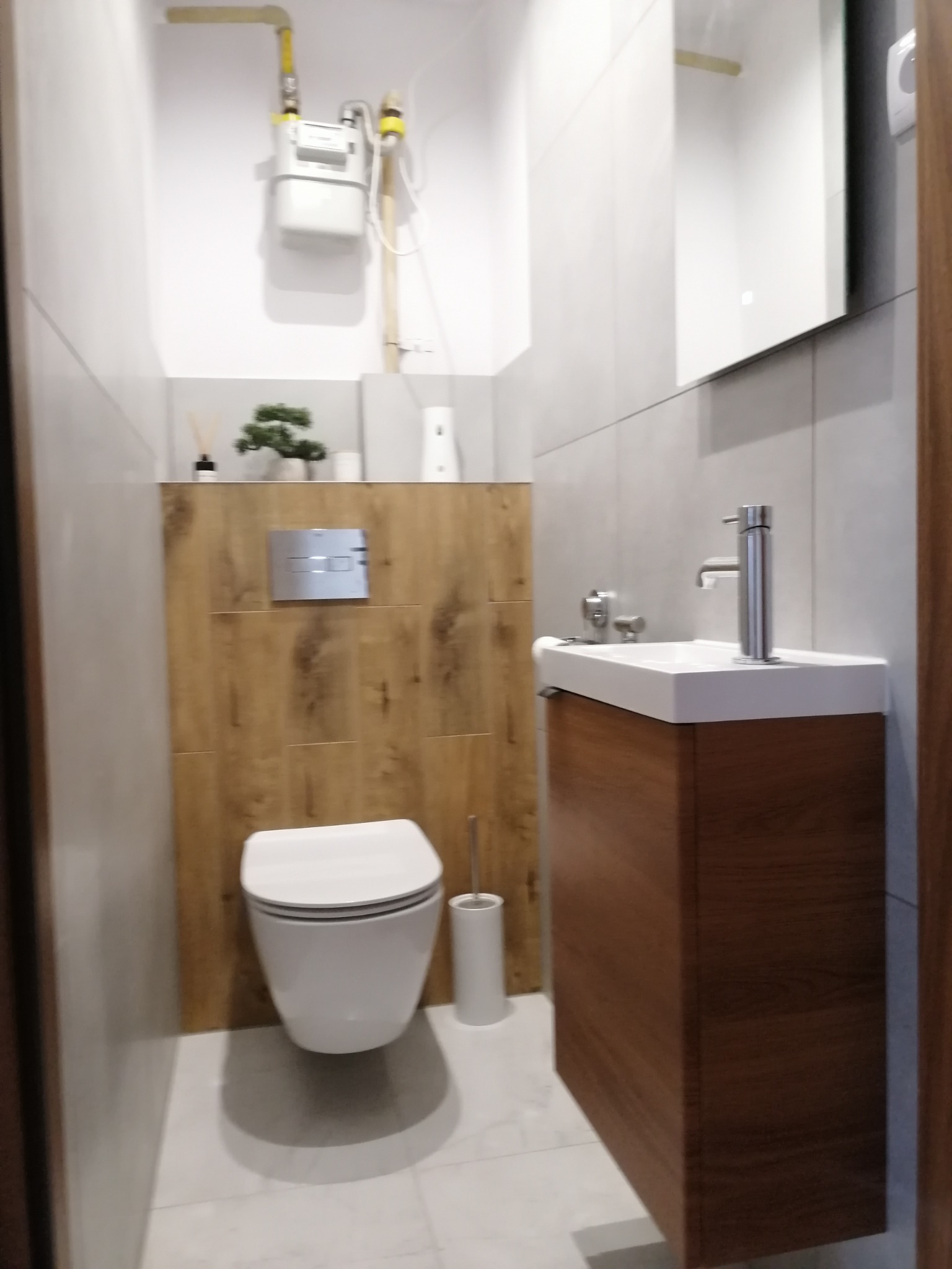 Nowoczesna łazienka z podwieszaną toaletą, umywalką nablatową na drewnianej szafce, szarymi płytkami i drewnianą ścianą za toaletą, na której zamontowano przycisk spłukujący.