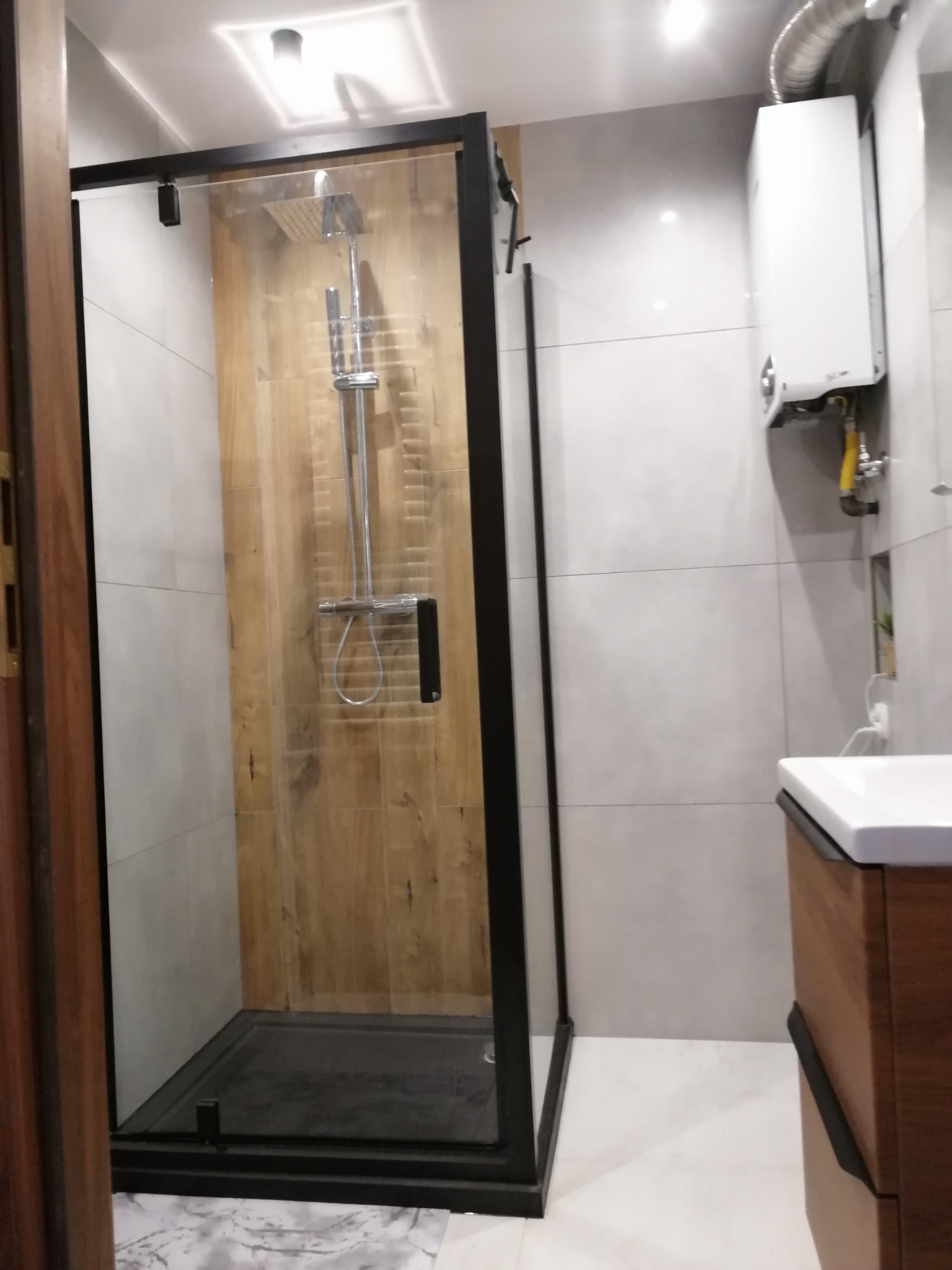 Nowoczesna kabina prysznicowa z czarną ramą i drewnopodobnym panelem ściennym, widoczny fragment umywalki i piec gazowy.