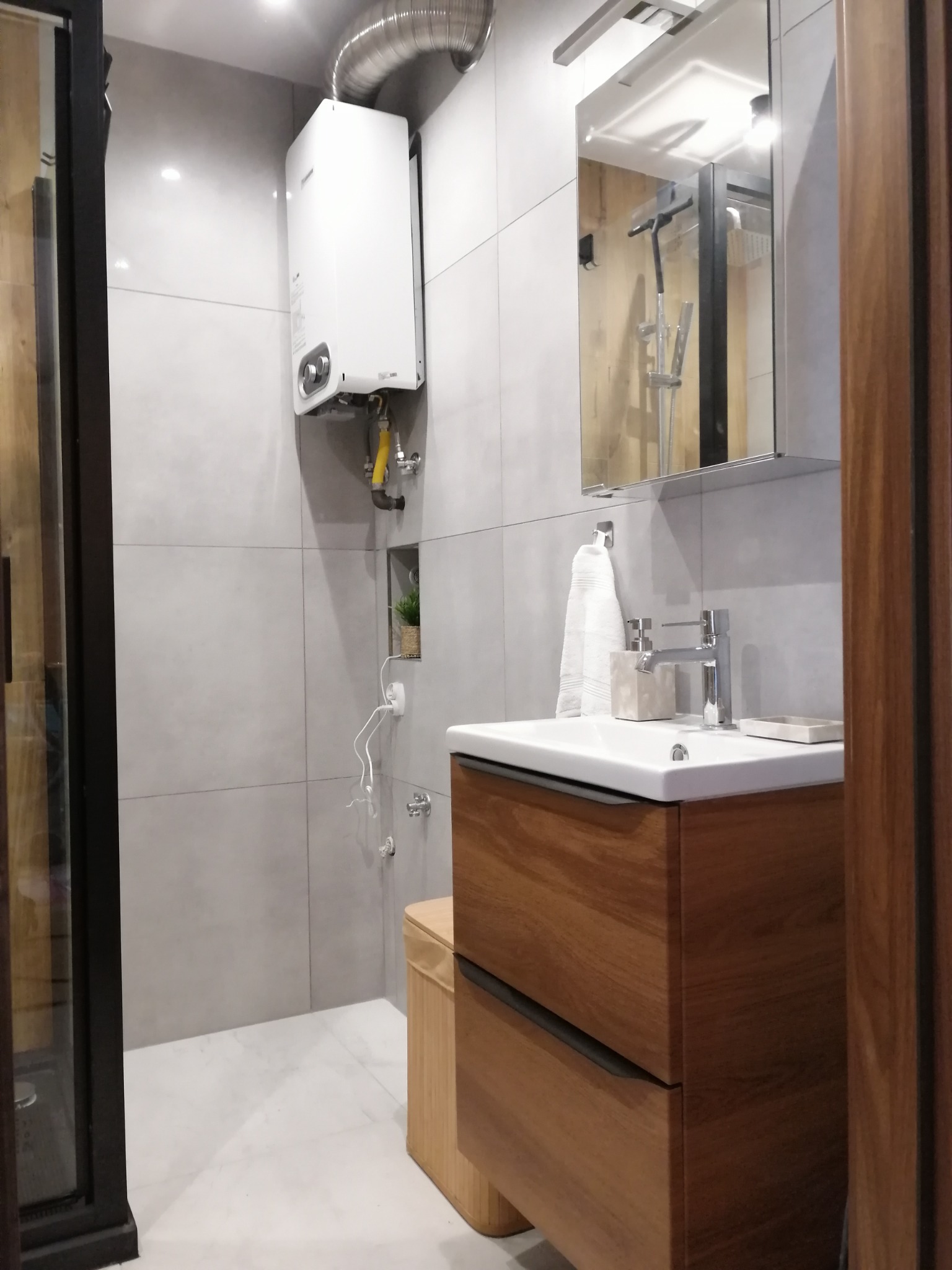 Nowoczesna łazienka z drewnianą szafką pod umywalką, grzejnikiem gazowym z rurą odprowadzającą spaliny oraz lustrem odbijającym kabinę prysznicową.