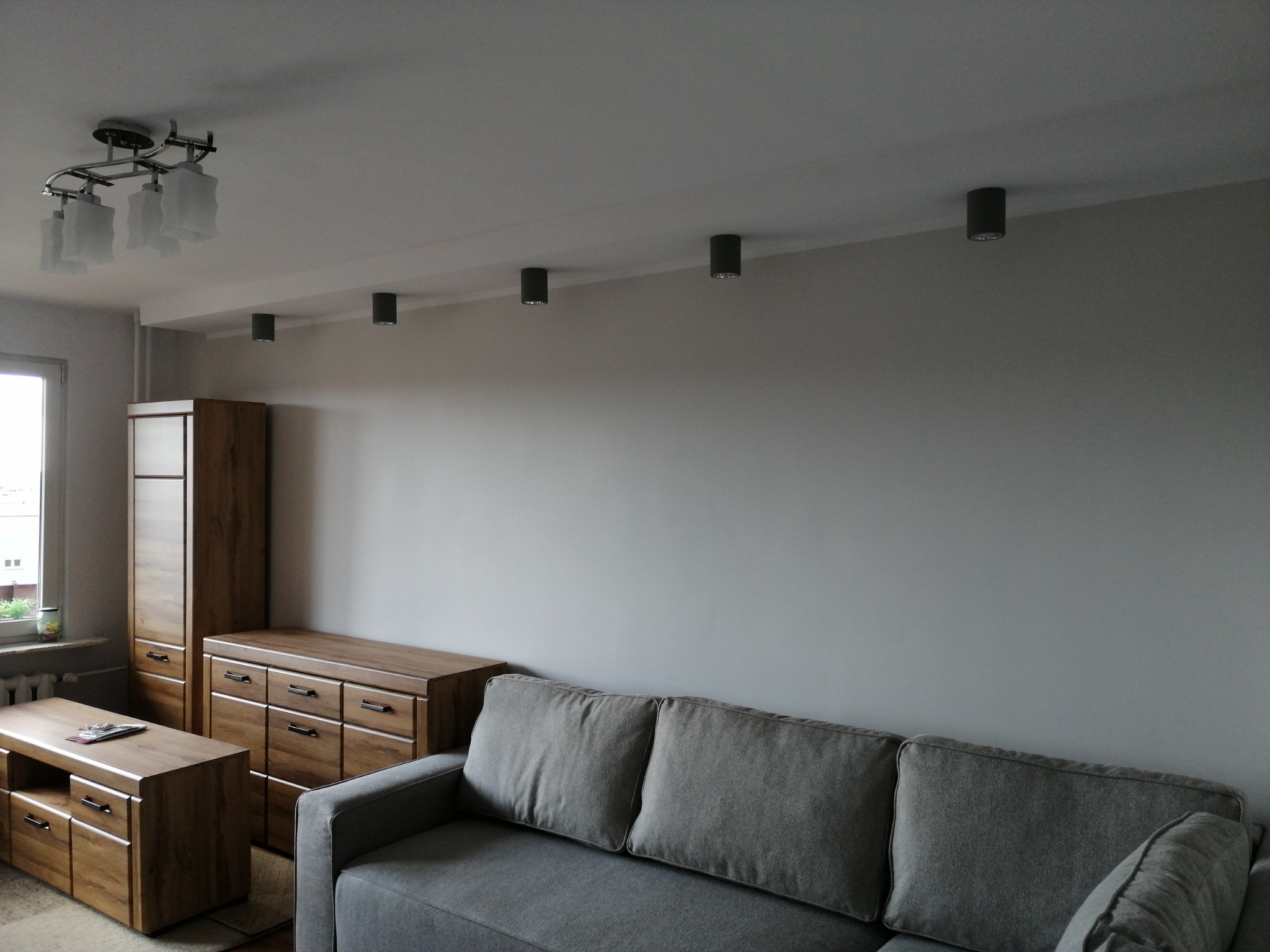 Szara sofa w salonie z drewnianymi meblami i oświetleniem sufitowym punktowym oraz lampą wiszącą.