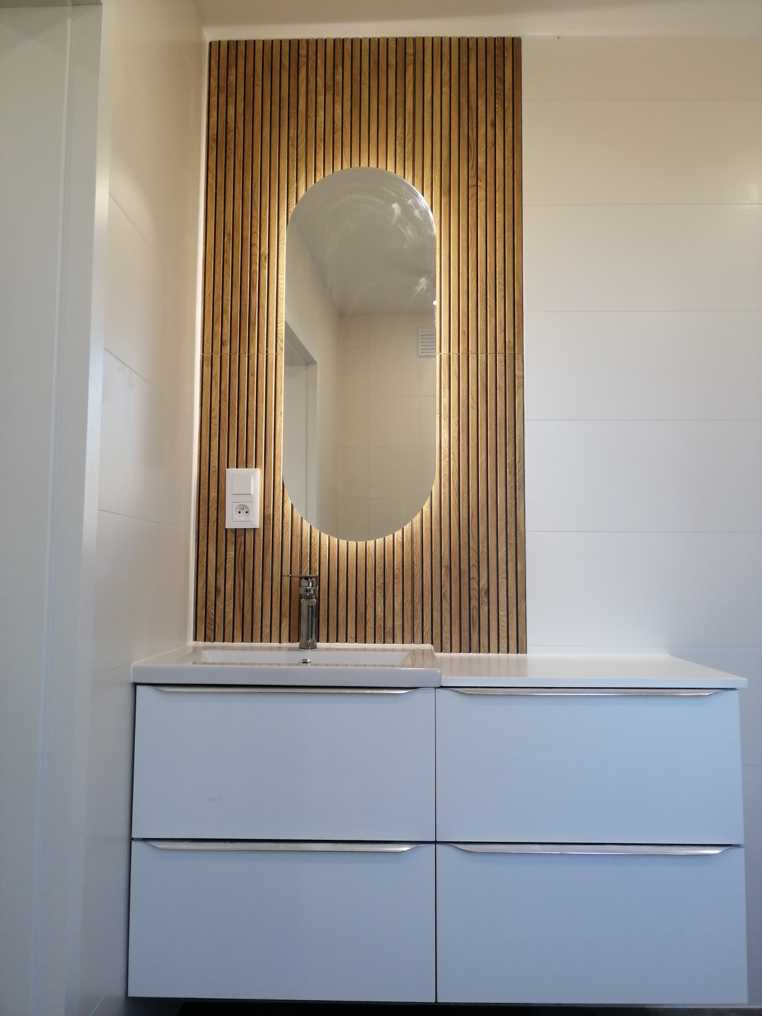 Nowoczesna łazienka: biała szafka z umywalką, podświetlane lustro w drewnianej ramie z lameli, białe płytki na ścianie i gniazdko elektryczne.