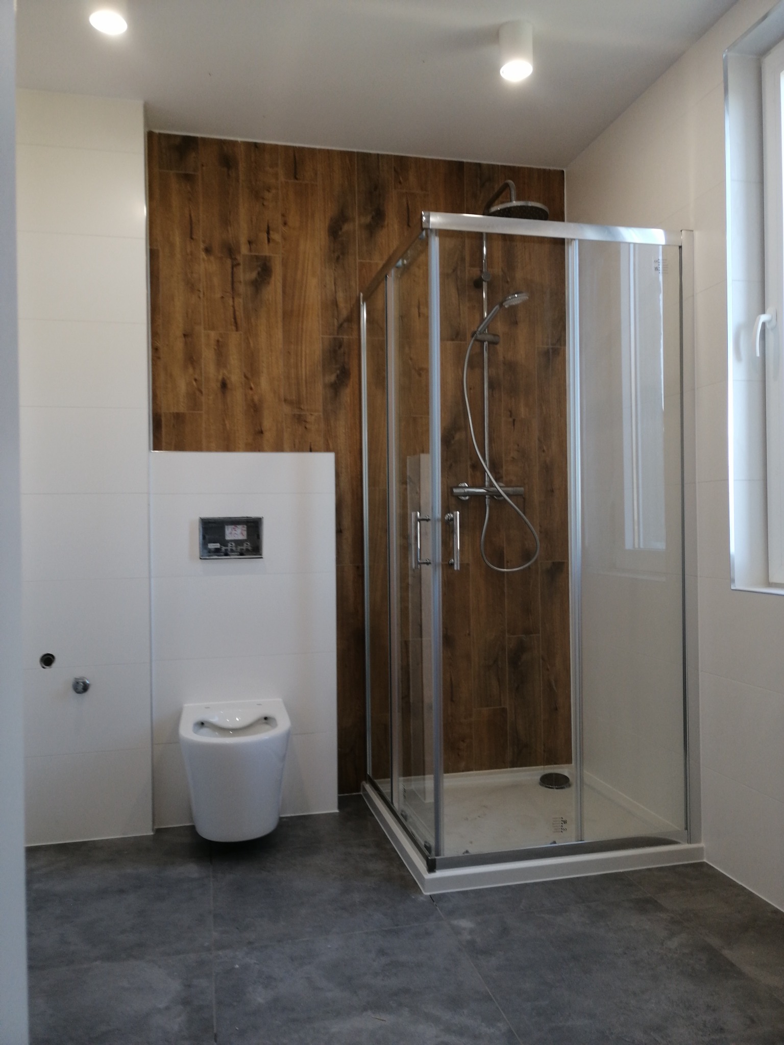 Nowoczesna łazienka po remoncie: kabina prysznicowa z drewnopodobnymi płytkami, podwieszana toaleta i szare płytki podłogowe. Minimalistyczny design.
