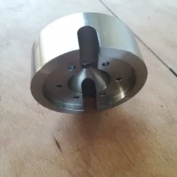 Precyzyjnie obrobiony, metalowy element cylindryczny z dwoma wycięciami i otworami montażowymi, leżący na drewnianej powierzchni.