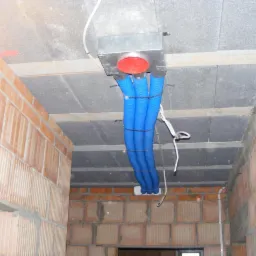 Cztery niebieskie, karbowane rury doprowadzone do metalowej skrzynki z czerwonym wlotem, zamontowanej w surowym, betonowym stropie budynku w trakcie budowy, widoczne ściany z cegły.