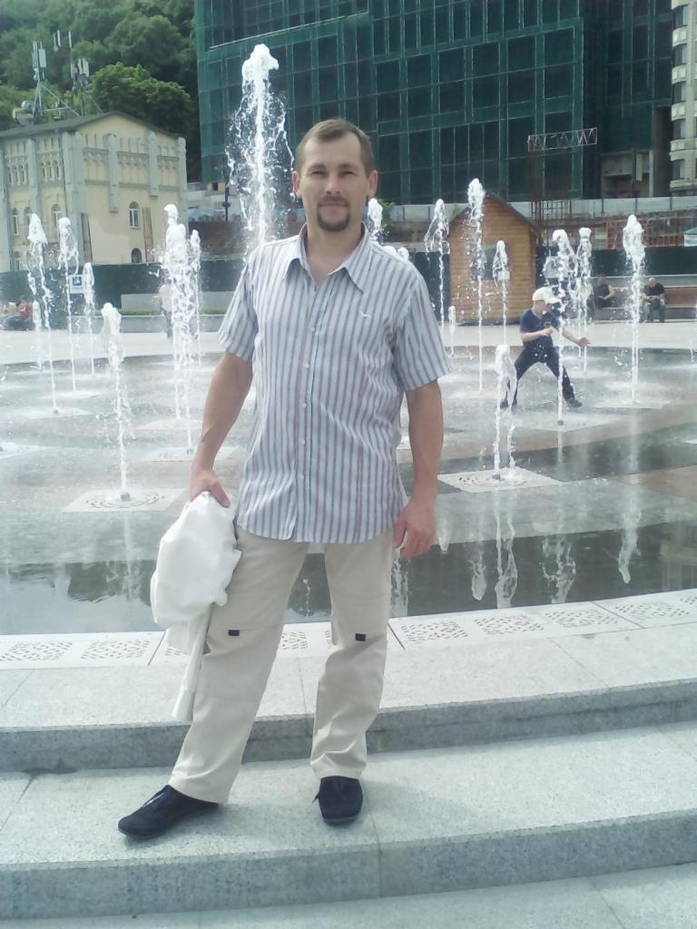 Mężczyzna w koszuli w paski i beżowych spodniach stoi na tle fontanny miejskiej z tryskającą wodą.