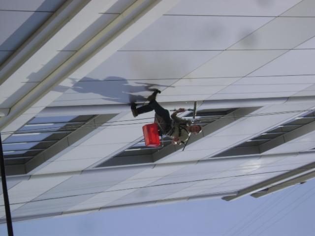 Pracownik na linach czyści okna wysokiego biurowca, widoczny z dołu pod kątem, z czerwonym wiadrem i narzędziami.