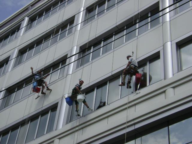 Trzech alpinistów na linach czyści okna wysokiego biurowca, jeden z nich z uniesioną ręką, widoczne wiadra i sprzęt do mycia.