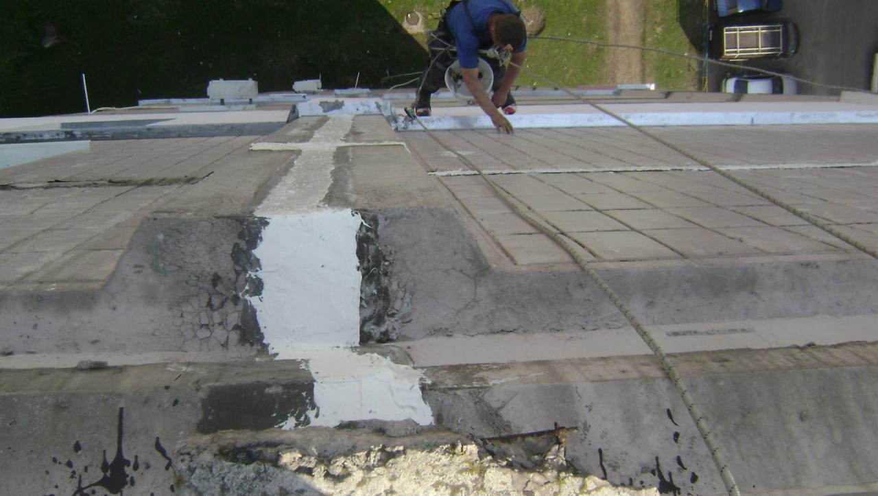 Pracownik zabezpieczony linami wykonuje naprawę betonowego dachu, wypełniając ubytki białą masą uszczelniającą.