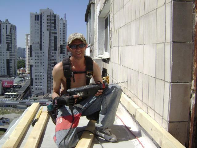 Mężczyzna w okularach i roboczych rękawicach wierci wiertarką w ścianie budynku z płytek, widok na miasto w tle.