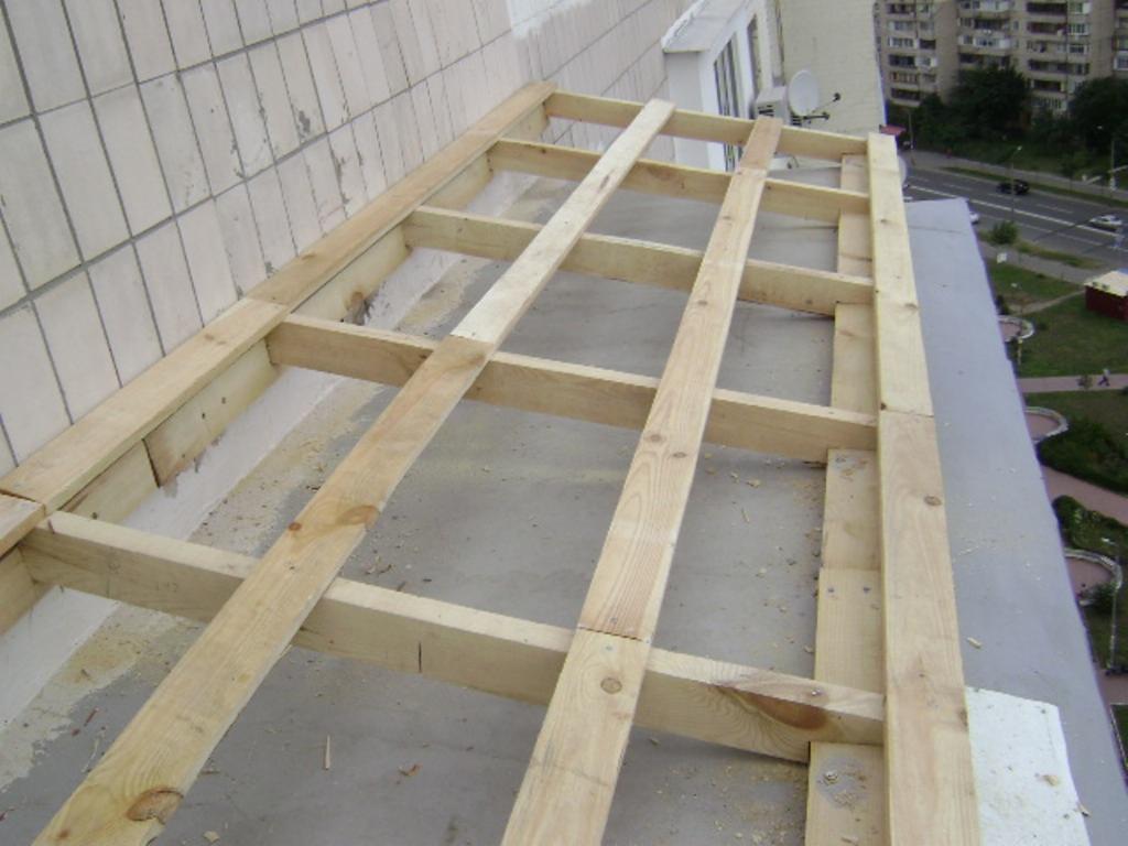 Drewniana konstrukcja dachu płaskiego w trakcie budowy, widok z góry na surową konstrukcję z belek, w tle budynki mieszkalne i zieleń.