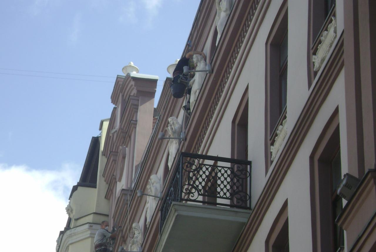 Dwóch pracowników na rusztowaniu linowym podczas renowacji elewacji kamienicy z balkonem i rzeźbami, widok od dołu.