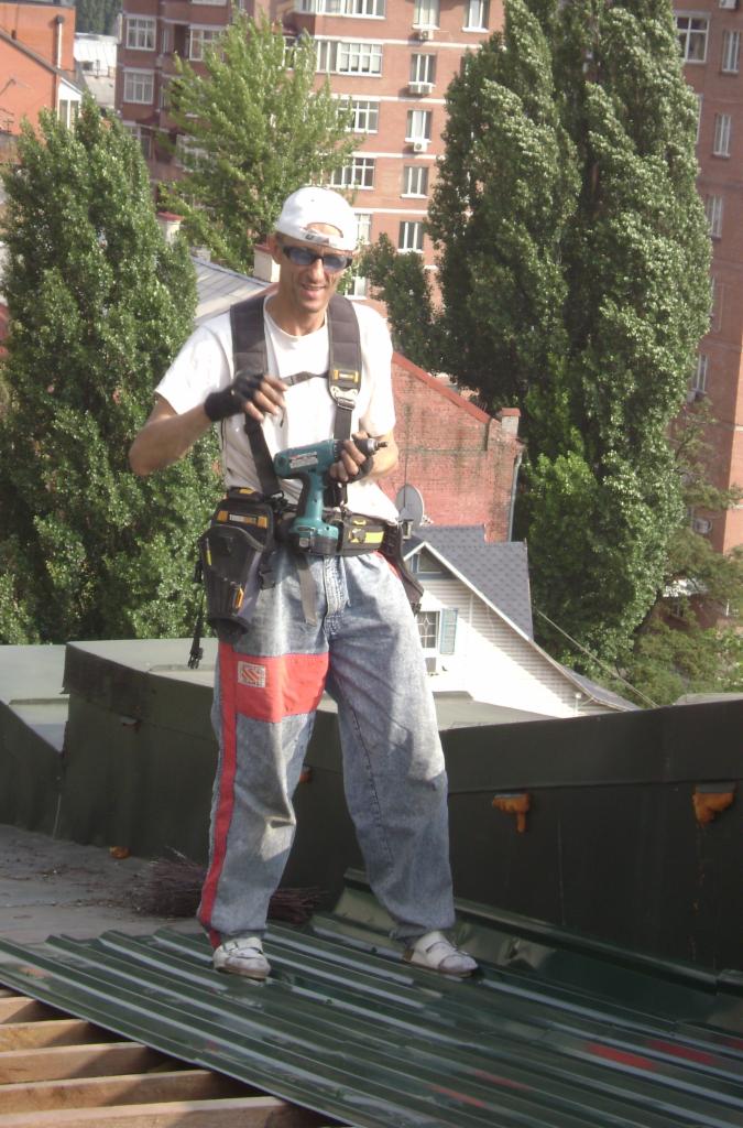 Pracownik w okularach, z wiertarko-wkrętarką, w szelkach i spodniach roboczych z czerwonymi wstawkami, stoi na blaszanym dachu podczas montażu.