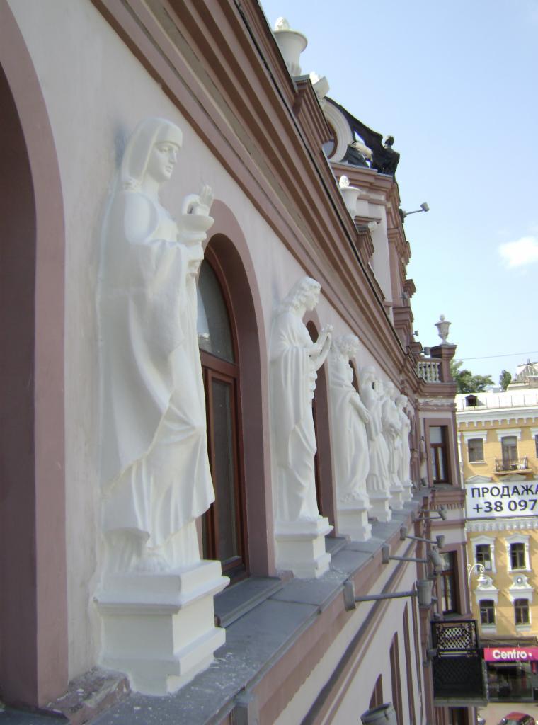 Fasada budynku z rzędem białych, klasycznych rzeźb kobiecych, każda z nich umieszczona na cokole, widoczne okna z brązowymi ramami i fragmenty budynku sąsiadującego.