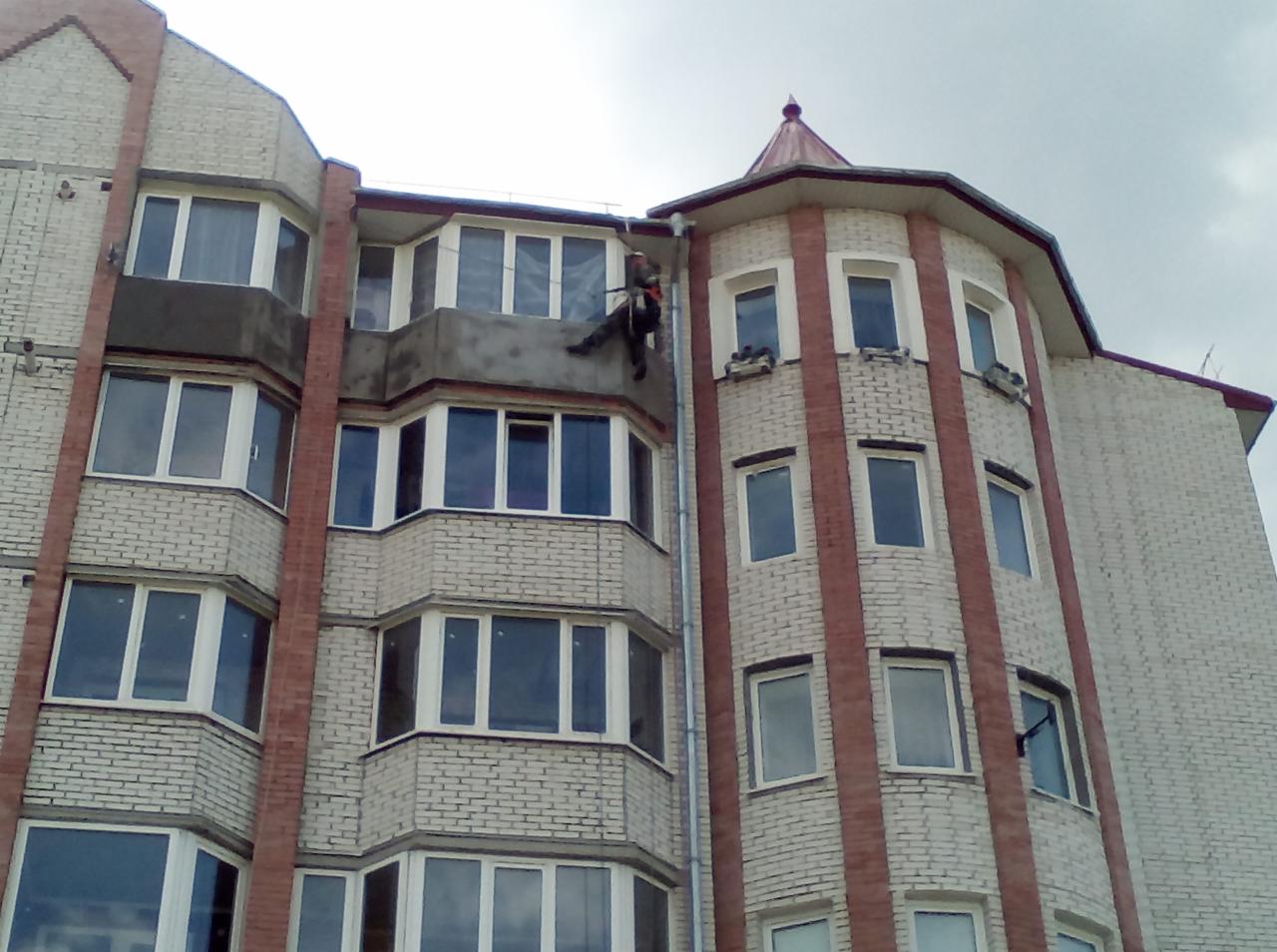 Pracownik na linie zabezpieczonej na dachu bloku mieszkalnego, prawdopodobnie podczas prac remontowych lub naprawczych na wysokości.