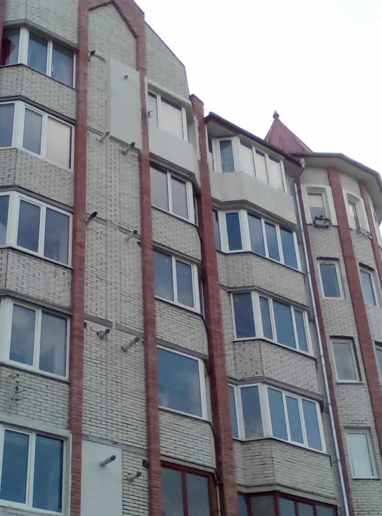 Wielopiętrowy budynek mieszkalny z jasnej cegły z ciemnoczerwonymi słupami, widok z dołu na fragment elewacji i okna.