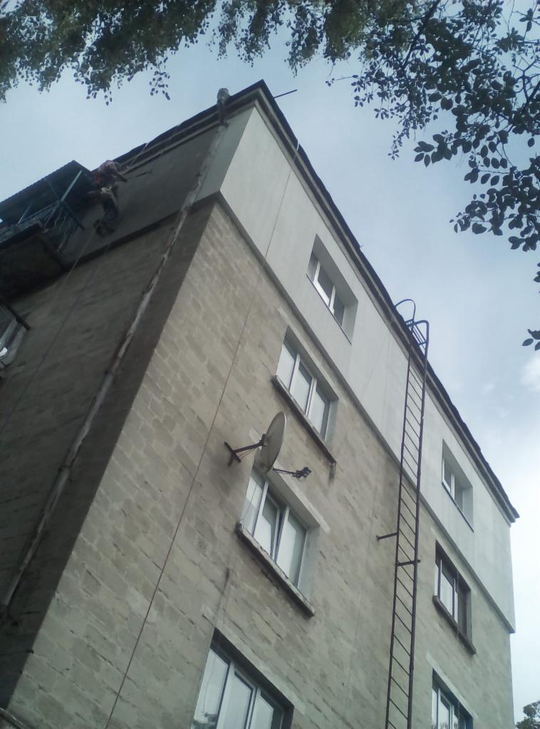 Pracownik na wysokościach, zabezpieczony linami, wykonuje prace wykończeniowe na elewacji bloku mieszkalnego, widoczna drabina, antena satelitarna i balkon.