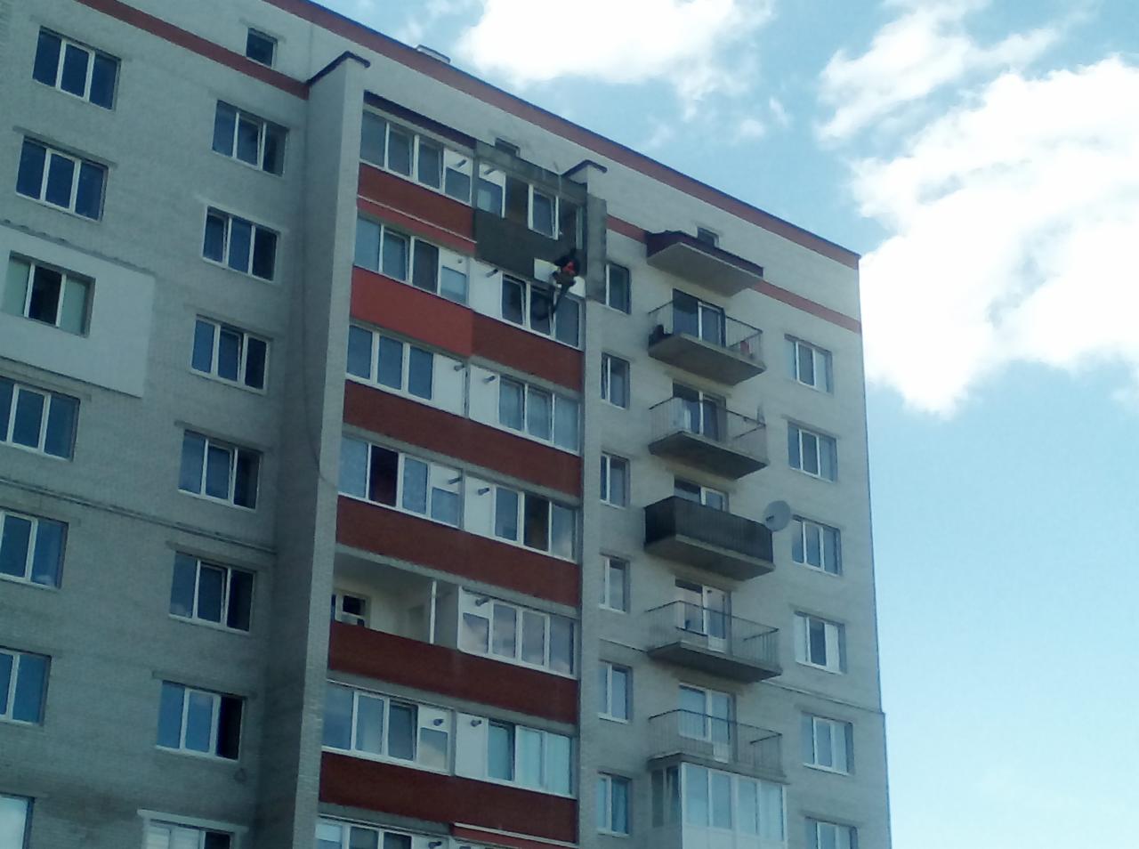 Pracownik na rusztowaniu przy ocieplaniu ściany bloku mieszkalnego z balkonami, widoczne okna i fragment błękitnego nieba.