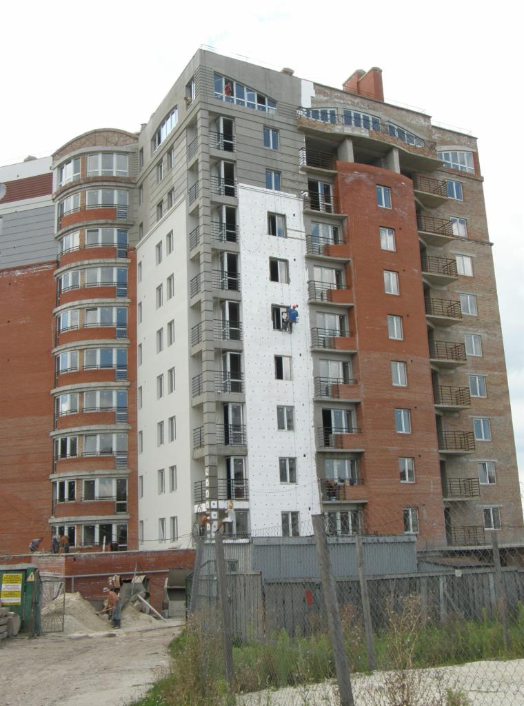 Wielopiętrowy budynek w trakcie ocieplania elewacji, robotnicy na rusztowaniu mocują białe płyty izolacyjne, widoczne fragmenty ceglanej elewacji i balkony z metalowymi barierkami.