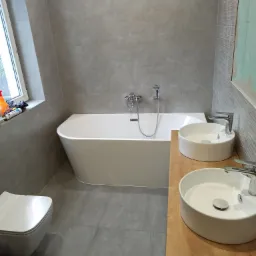 Nowoczesna łazienka z wolnostojącą wanną, dwiema okrągłymi umywalkami nablatowymi na drewnianym blacie i wiszącą toaletą, wykończona szarą płytką ceramiczną.