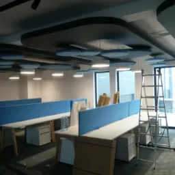 Nowoczesne biuro z białymi biurkami, niebieskimi przegrodami, podwieszanymi sufitami z okrągłymi panelami akustycznymi i drabiną opartą o okno.