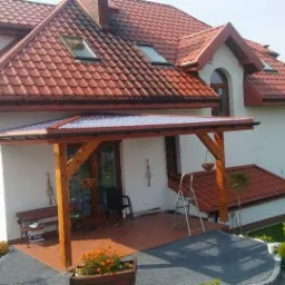 Dom z czerwoną dachówką i drewnianą konstrukcją zadaszenia tarasu z przezroczystym pokryciem, widok z ogrodu z kwiatami.