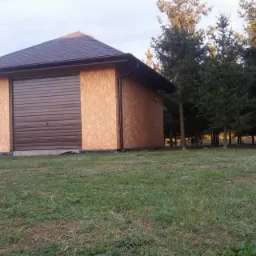 Niewielka, brązowa altana z dachem pokrytym gontem bitumicznym i ścianami z płyty OSB, zamykana na brązową bramę garażową, otoczona trawnikiem i drzewami iglastymi.