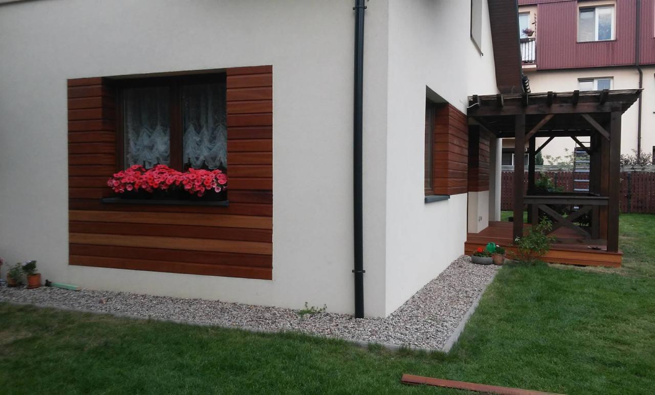 Elewacja domu z drewnianą okładziną okienną, skrzynką z czerwonymi kwiatami i drewnianą altaną na tarasie, widok z trawnika.