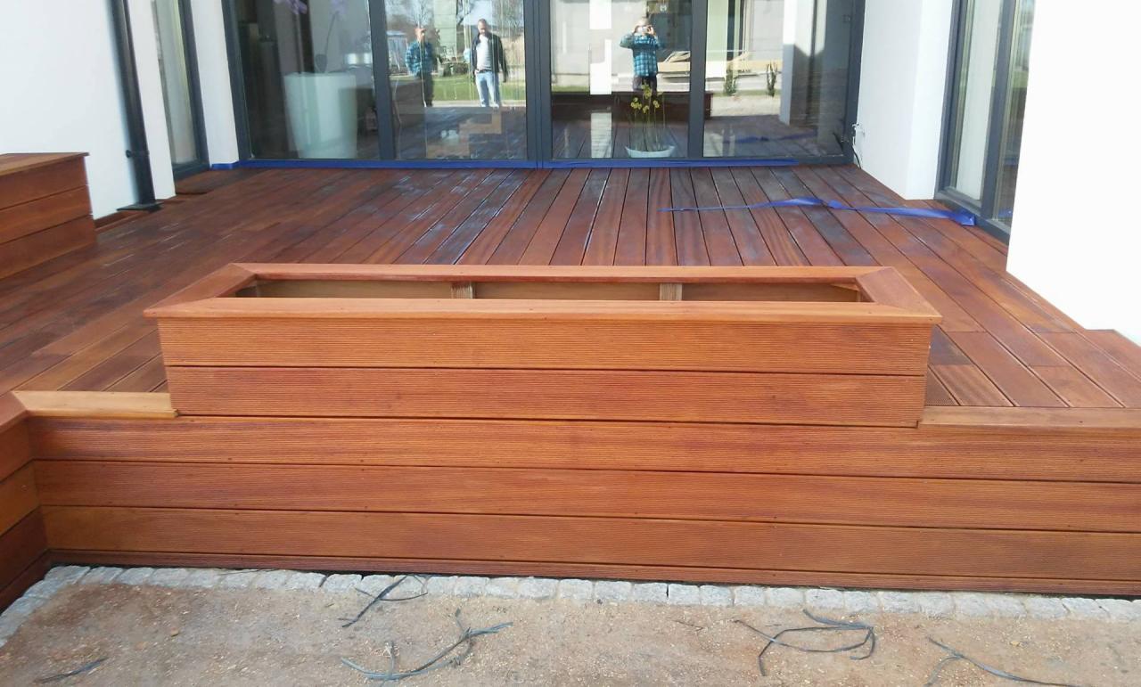 Nowoczesny taras z drewna egzotycznego, z wbudowaną donicą, na tle dużych okien przesuwnych.