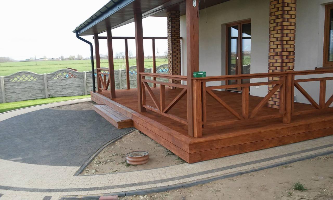 Zadaszony taras drewniany z brązowymi balustradami i ceglanymi kolumnami, widok z poziomu gruntu na tle pola i betonowego ogrodzenia.