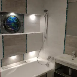 Wykończona łazienka z wanną obudowaną płytkami w odcieniach szarości i beżu, z dekoracyjnym okrągłym panelem z motywem podwodnego świata i ukrytym oświetleniem LED nad wanną.