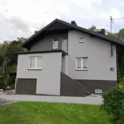 Dwukondygnacyjny dom jednorodzinny z szarą elewacją, czarnym dachem i podmurówką wykończoną ciemną, teksturowaną płytką, widok z trawnika, dwa białe wiadra po farbie w prawym dolnym rogu.