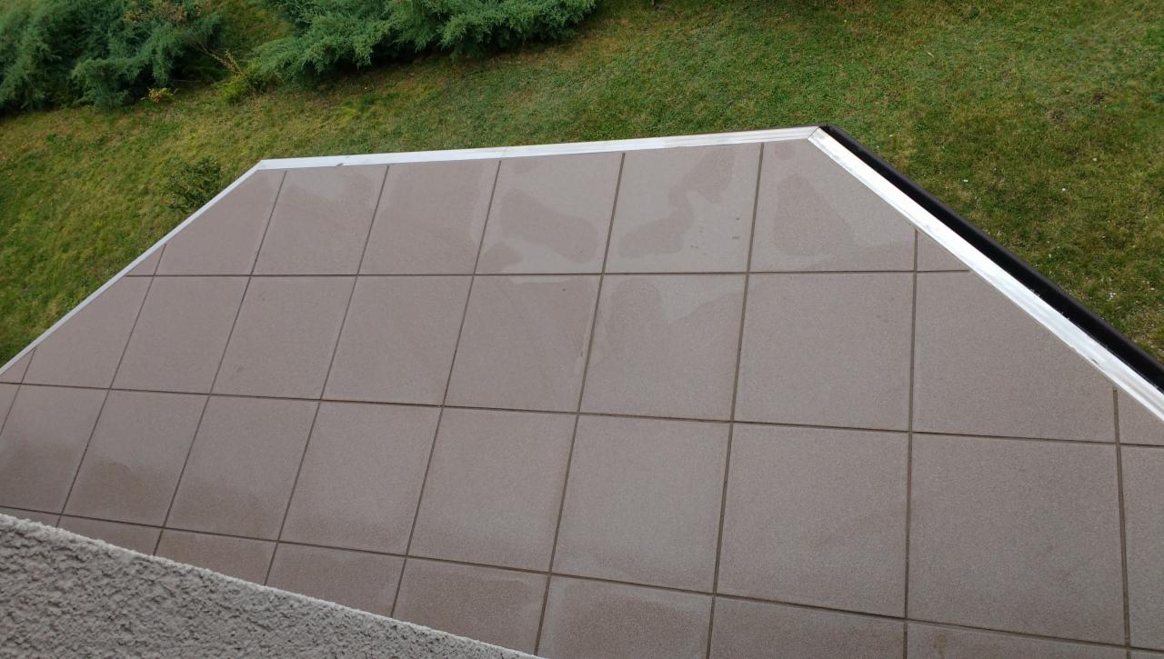 Ułożone, kwadratowe, beżowe płytki na tarasie z aluminiowym obramowaniem, widok z góry, w tle trawnik i krzewy.