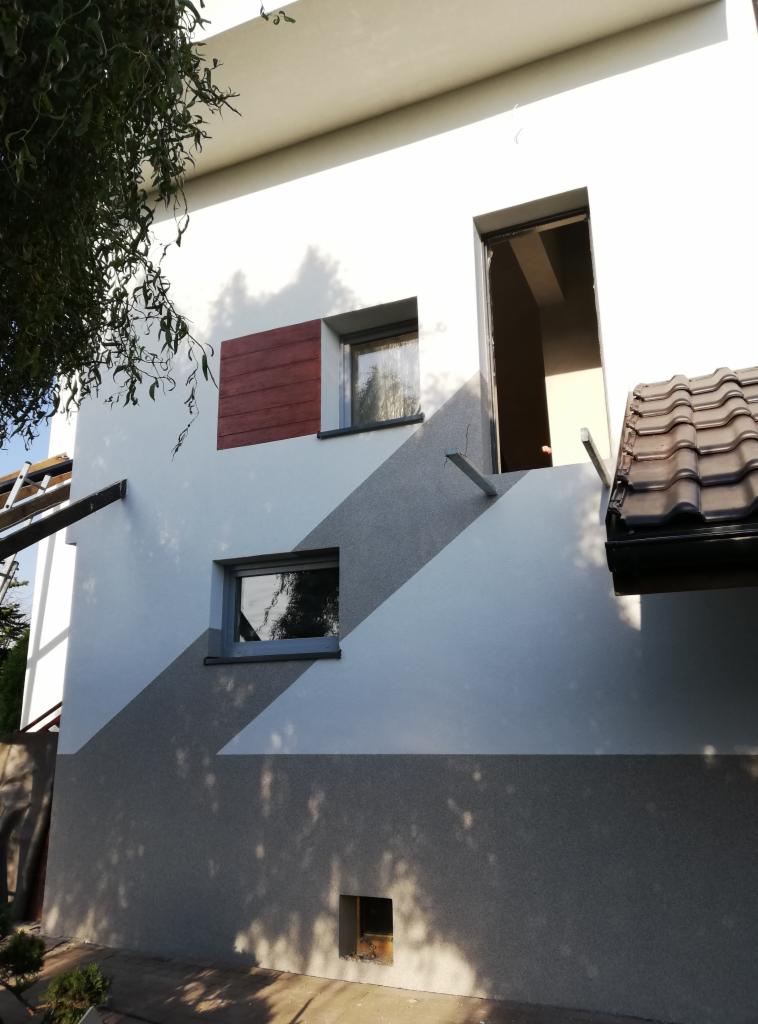 Elewacja domu z szarymi pasami, oknami i fragmentem drewnianej okładziny, widok z dołu na częściowo oświetloną fasadę.